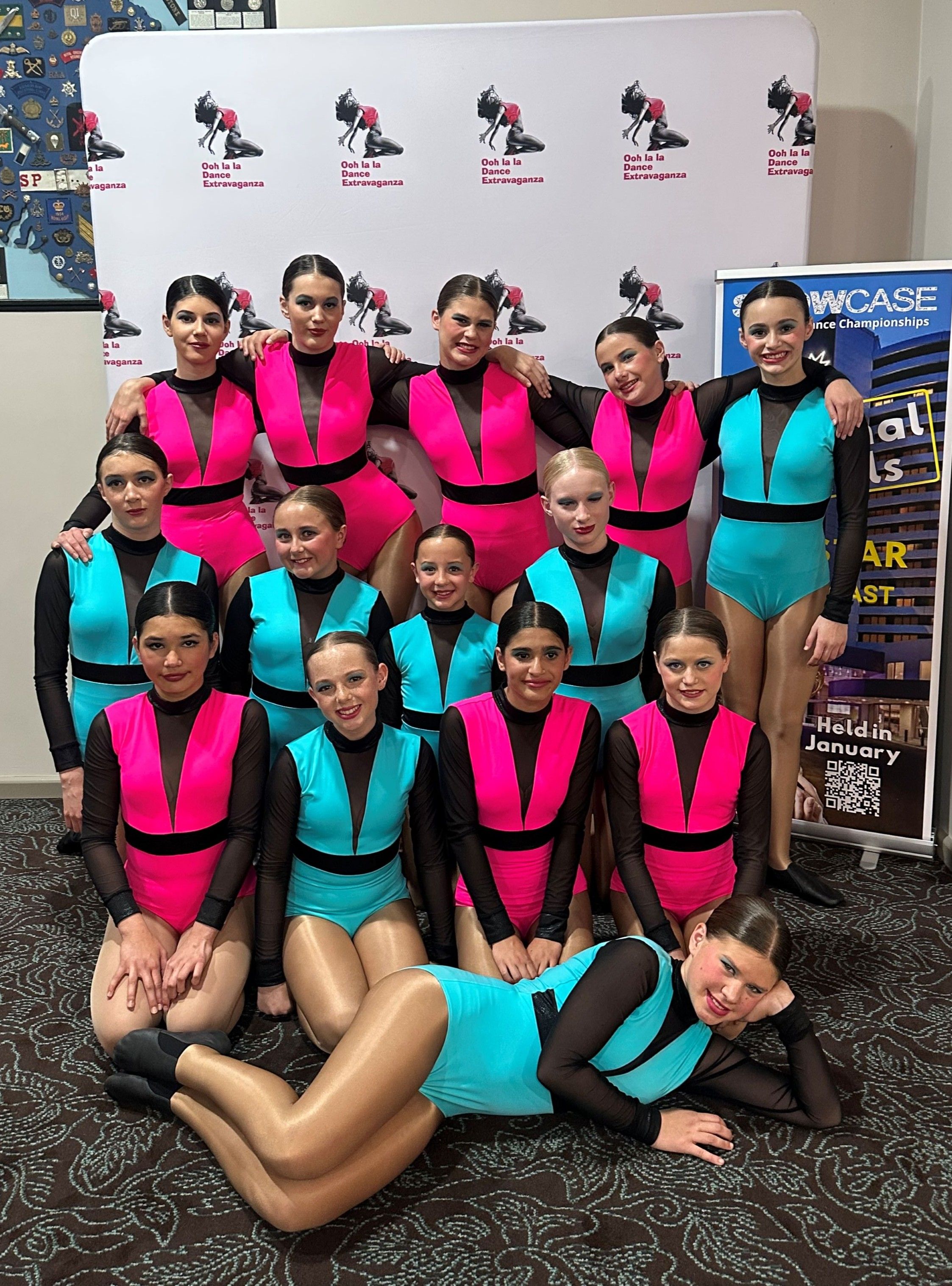 Fourteen teenage dancers pose for a group photograph in pink, blue and black costumes.