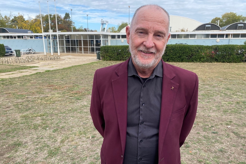 A man with grey hair and a beard stands on grass outside an Olympic pool.