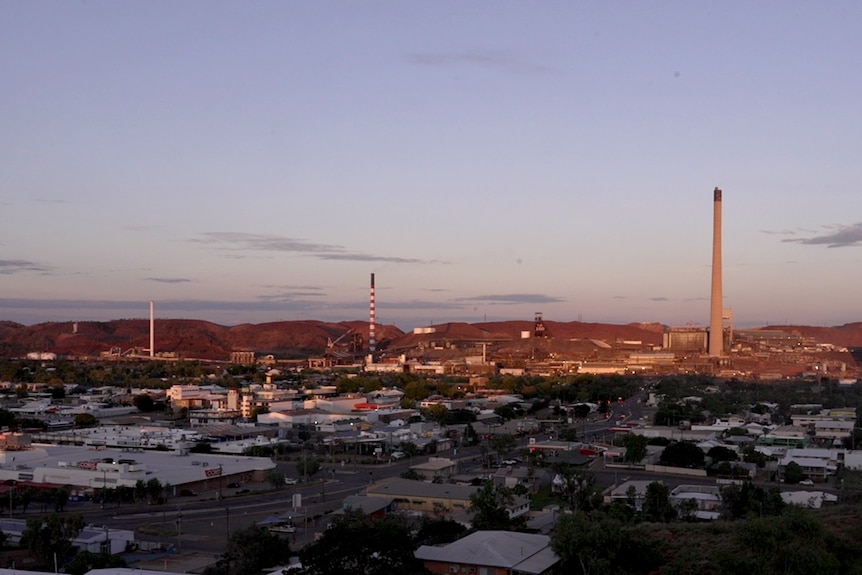 The roofs of buildings and three smelter stacks from the Mount Isa mine against a blueish purple sky.
