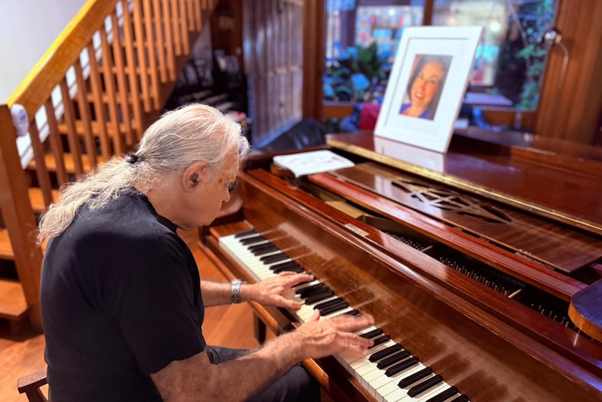 Vincenzo sits at a piano in a lounge room, a photo of his wife on top of the piano