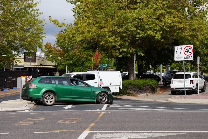 A crashed green Holden Commodore blocks a road in front of a shopping precinct.