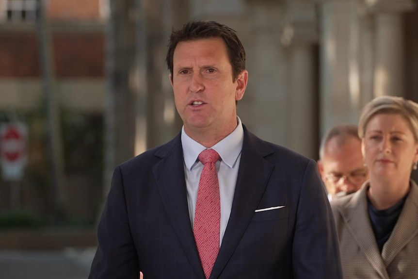 A man in a suit and pink tie speaking to media with a blonde woman standing behind him.