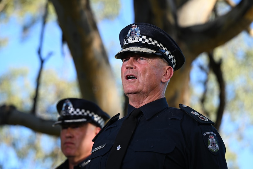A uniformed policeman stands speaking beneath a large tree.