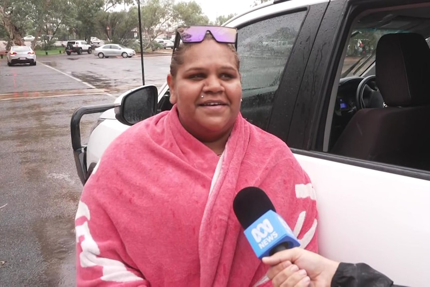An Aboriginal woman standing by white 4wd vehicle, pink warm jumper on, brown hair tied back, sunglasses resting on her head.