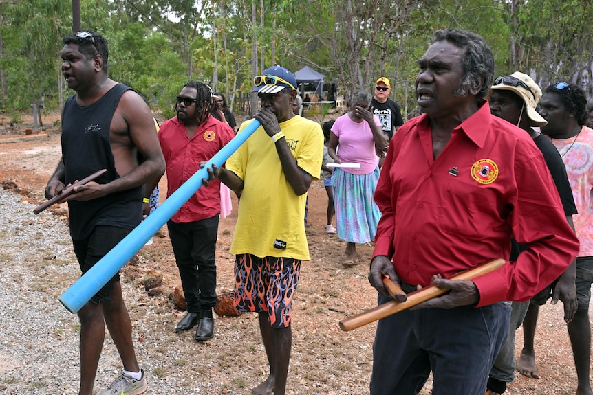A group of Indigenous Australians walk together outside. A man in front uses clap sticks, another plays a blue didge.