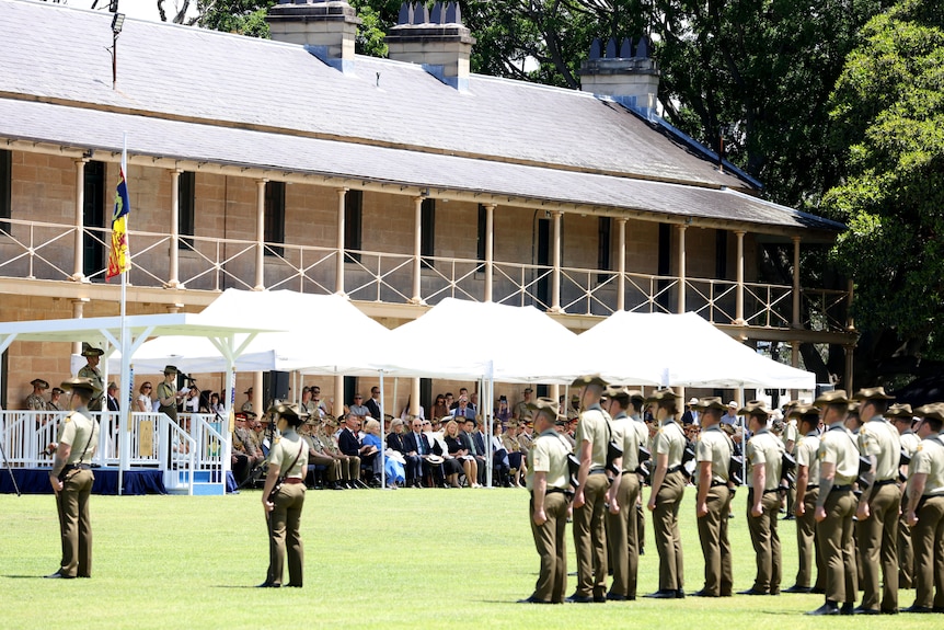A crowd of soldiers stand at attention in front of a large sandstone building. 