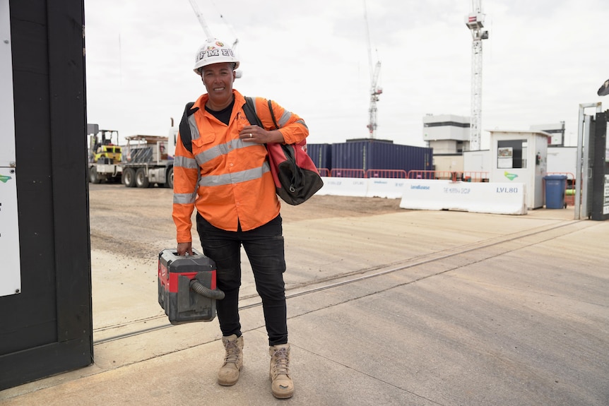 A woman wearing orange high-vis jacket and a white helmet walking on a street holding a tool bag.