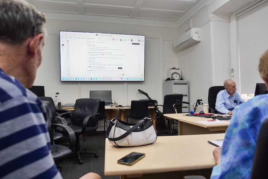 two elderly people watch a projector screen at the front of a classroom