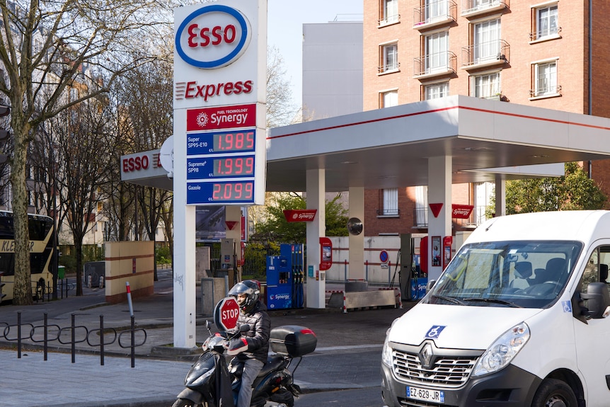 A motorcyclist in black clothing next to a white van on a road alongside a blue, white and red petrol station.