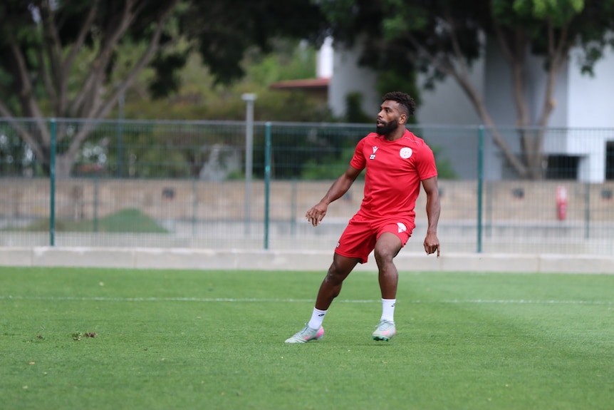 A Kanak man wearing a red football uniform and white football shoes and socks pushes off on his right leg on a soccer field.