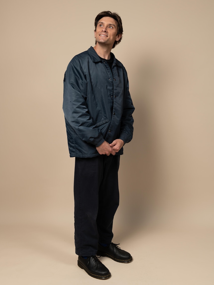 Sam Taunton, mid-30s, stands slightly to the side in a waterproof jacket, looking up and smiling brightly.