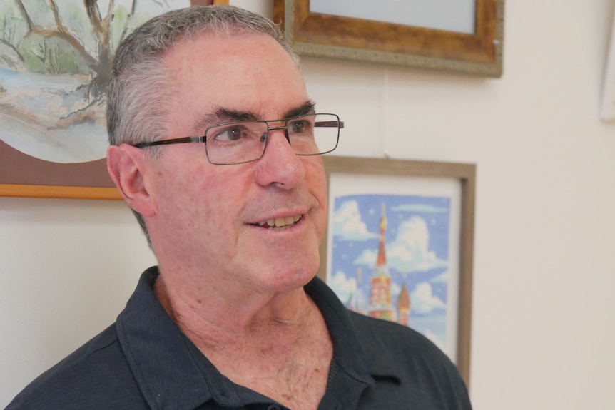 A middle-aged man with short grey hair stands in front of a wall with artworks on it.