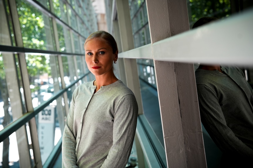 A woman wearing a grey cardigan with her hair in a pony tail looks seriously at the camera in a building corridor.