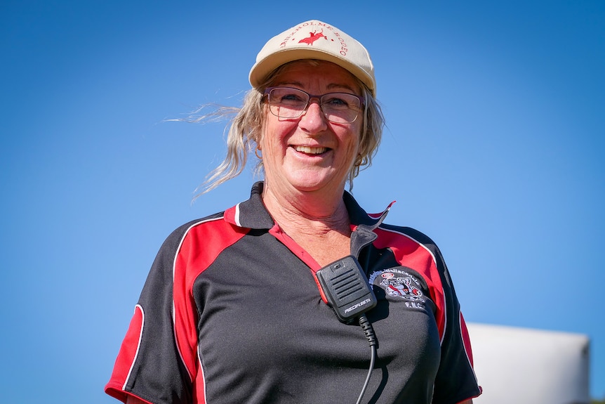 A woman wearing a rodeo branded cap, a football club polo and walkie talkie smiles at the camera. 