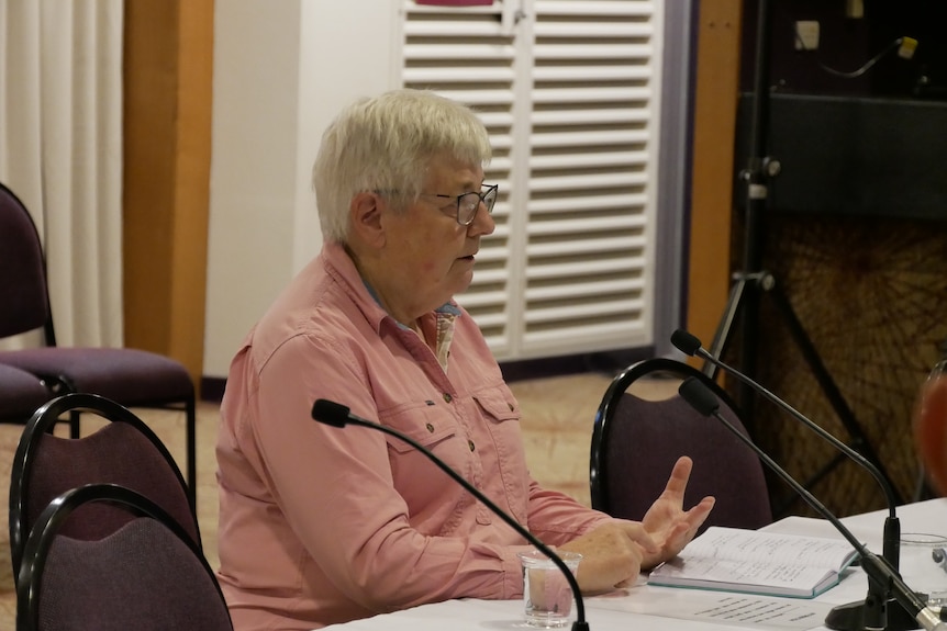 An older woman sits at a table in a conference room.