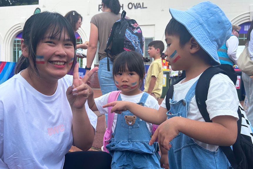 A woman with blue face paint makes a "peace sign" with two children next to her
