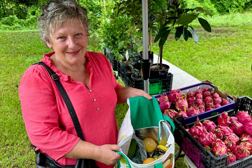A woman holds up a bag of produce in front of a farmers market stall.