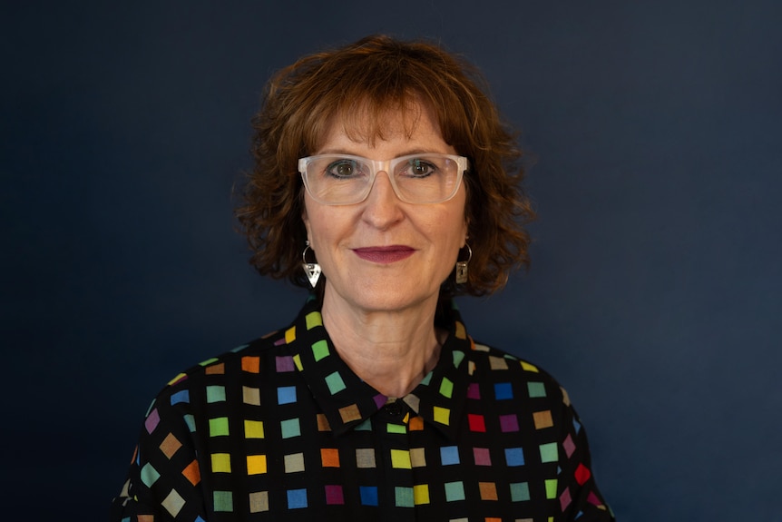 A headshot of a woman with glasses and a colourful shirt standing against a dark background. 
