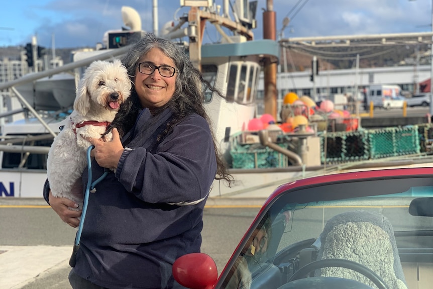 A woman with long dark hair and dark-framed spectacles poses for a photo on a harbour, next to a red car, holding her small dog