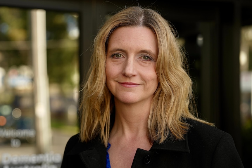 A woman with blond waving hair stands smiling at the camera in front of sliding doors