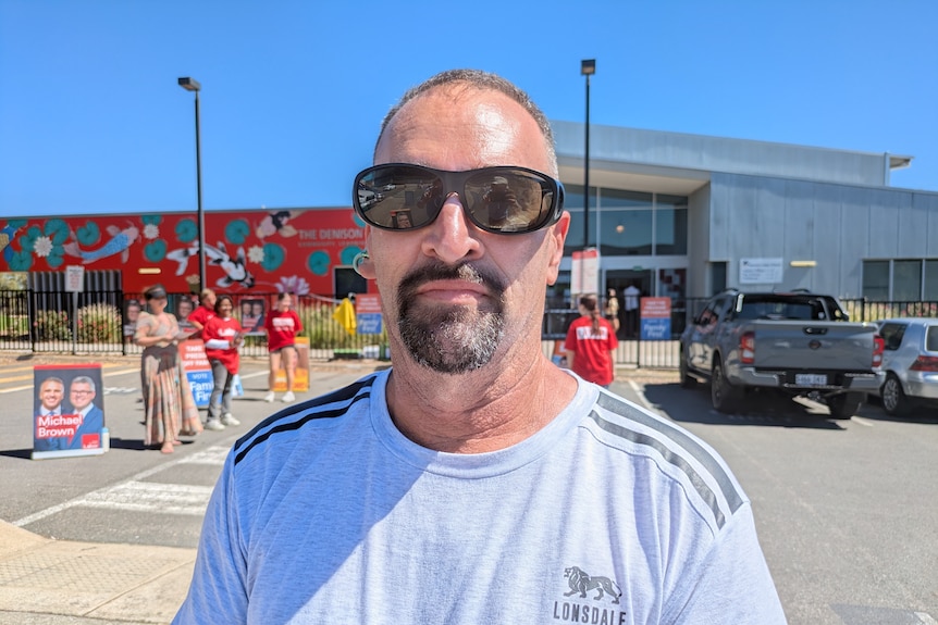 A man wearing sunglasses and a light grey top stands in front of a polling booth