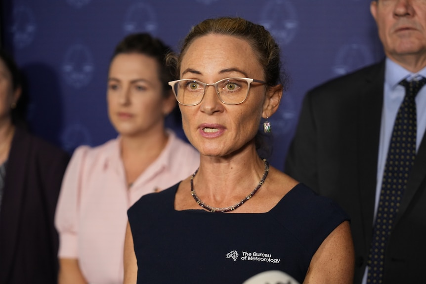 Shenagh Gamble wearing glasses and a sleeveless dress speaks to the media. 