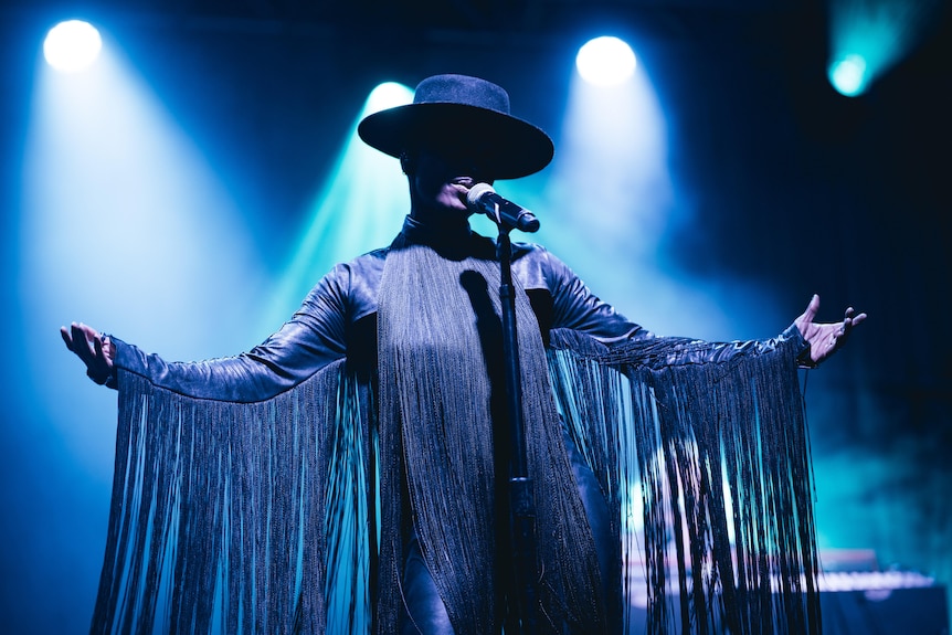 A singer in a face-obscuring wide-brimmed hat holds their arms out, fabric falling from each sleeve.