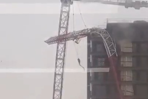 A crane bent over near a building, with a grey sky in the background.