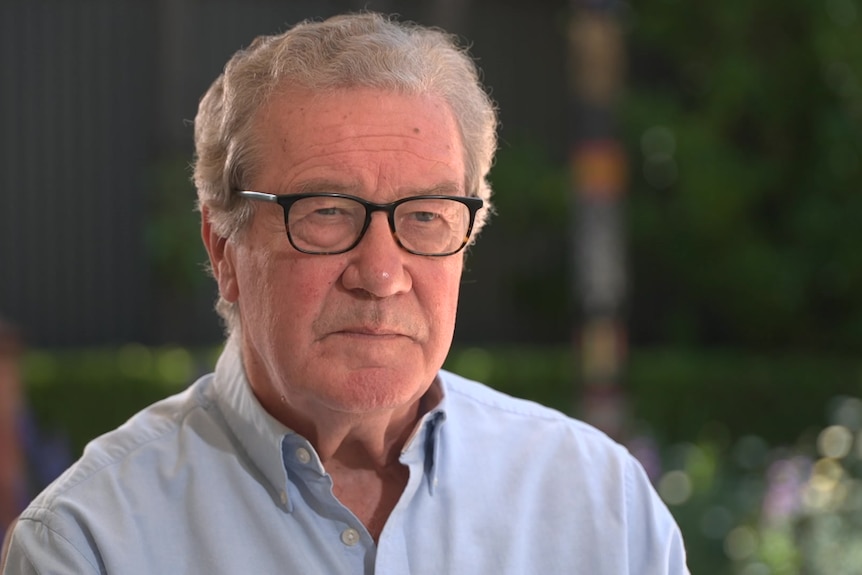 A man with short grey hair in a pale blue shirt wearing black-rimmed glasses sits looking off camera. 