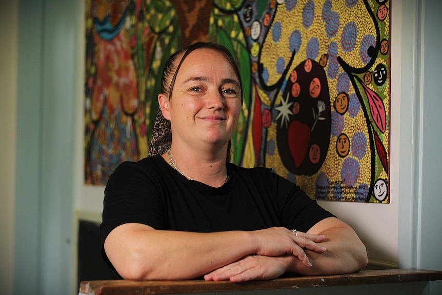 Woman with dark hair, arms folded, smiling in front of centre's Indigenous artwork.