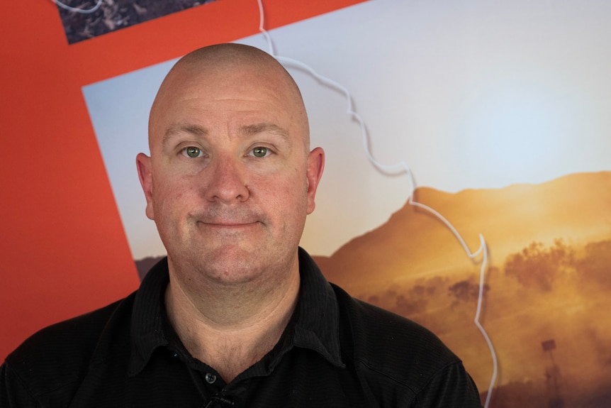A bald man smiles into camera, with a colourful backdrop behind him.