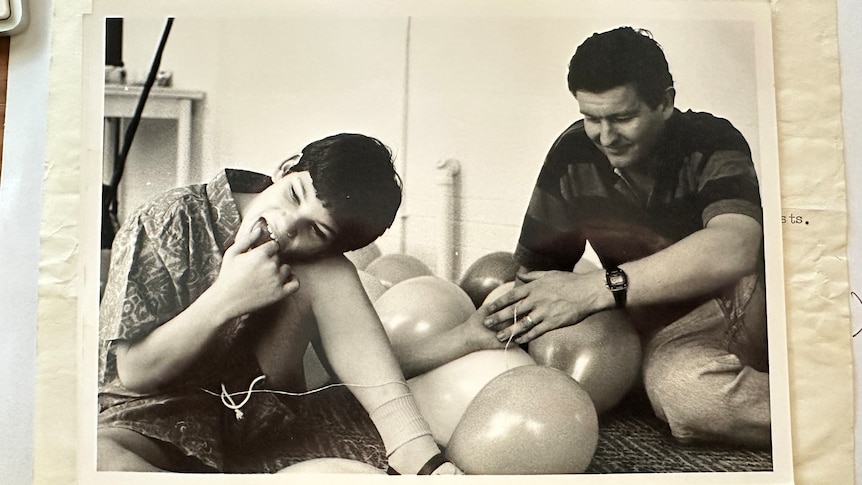 A 10-year-old boy with hand in his mouth sitting on floor with balloons next to him. An adult male sits beside him