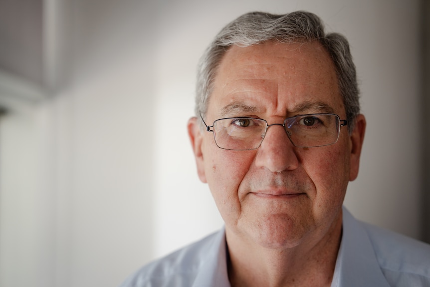 Close up shot of white man, gray short hair, looking directly into camera, light-framed glasses, neutral facial expression