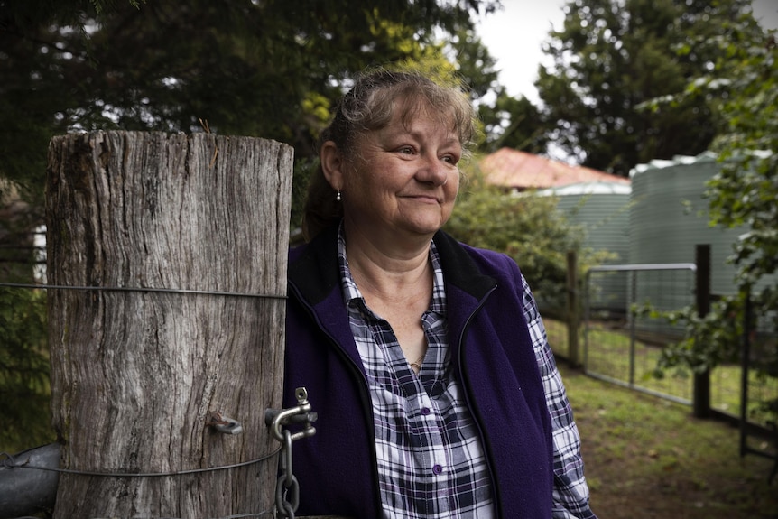 Woman with grey hair and checkered shirt leaning against wooden fence