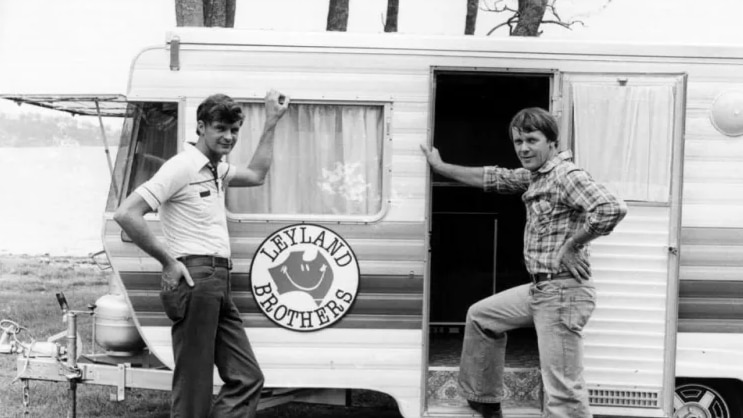Black and white imagery. Two men stand beside caravan. Their hands are leaning on the door frame and windows. Their hand on hip