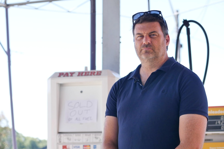 A dark-haired man in a polo shirt stands in front of a petrol bowser at a service station.