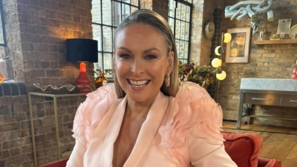 Smiling Mel in a peach jacket with flounces on shoulders, hair slicked back, stands in a room with brick walls, lamps.