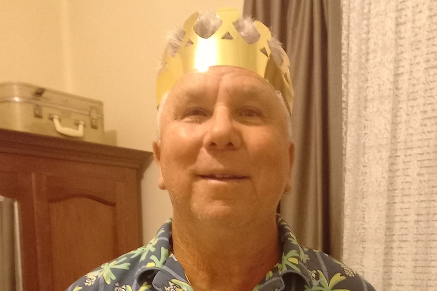 A man smiles while wearing a gold paper crown in front of a timber cabinet and a window with curtains