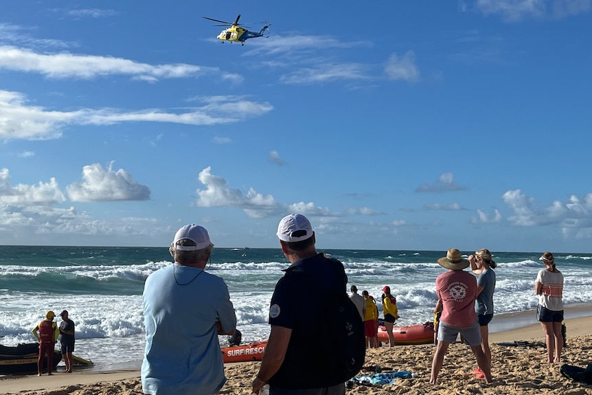 A helicopter flies over the ocean while people watch from a beach.