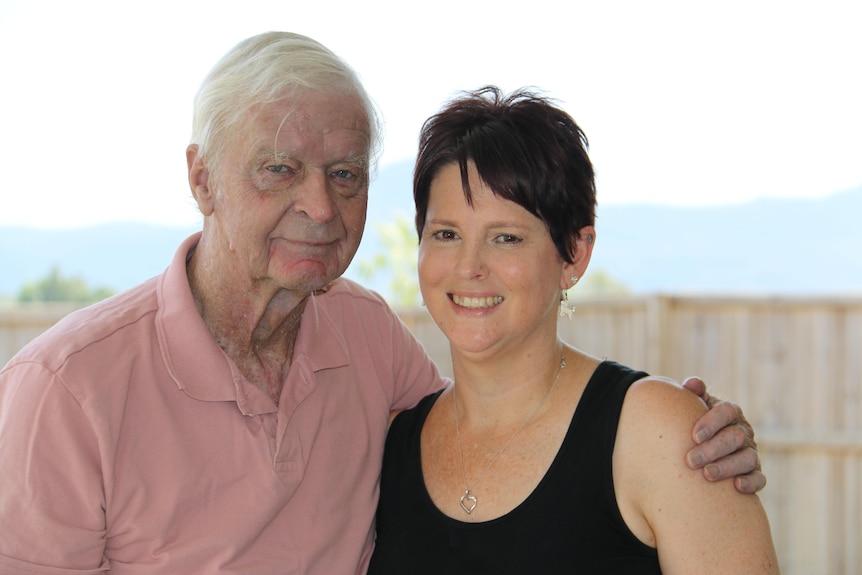 A man in his early 80s wearing a collared shirt has his arm around his adult daughter, she wears black singlet and short hair