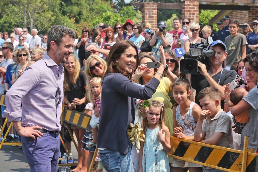 A bearded man with grey hair smiles as he watches a dark-haired woman wave to a crowd on a cordoned off city street.