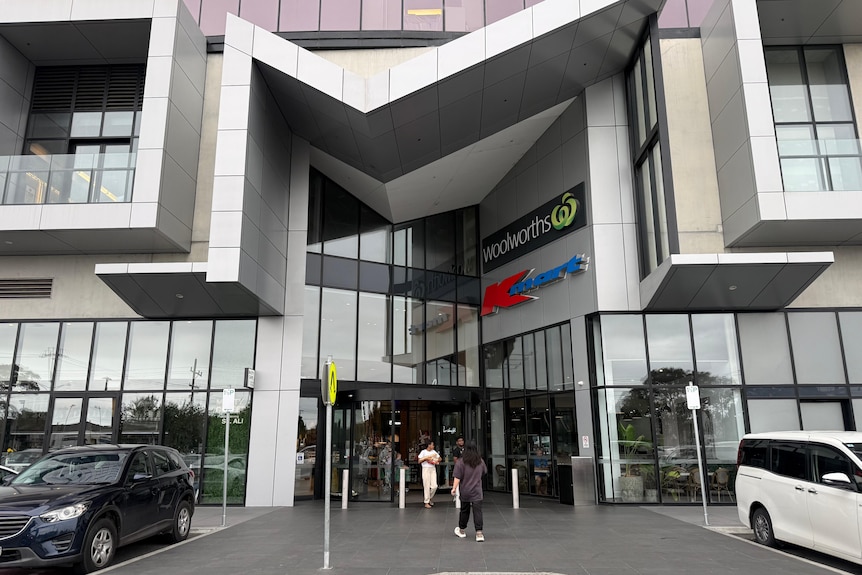 Two people walk outside the glass exterior of a multi-storey building that has a "Woolworths" and a "K Mart" sign on it.