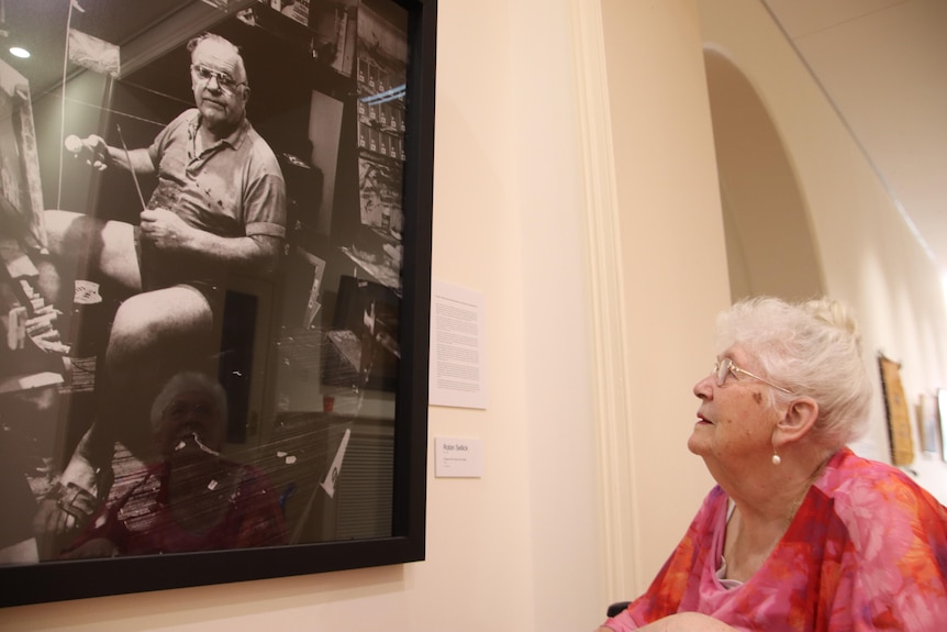 An old woman in a floral top and glasses looks at a large black and white photo of artist Pro Hart hanging on a wall.