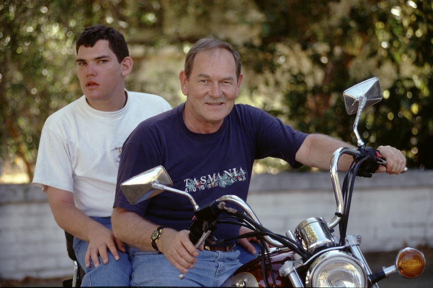 A young man in a white T-shirt sits on the back of a motorcycle, his father wears a navy shirt sitting at front hands on handles