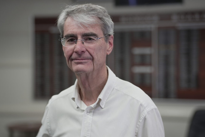 A middle-aged, grey-haired man in glasses stands in front  of a building.