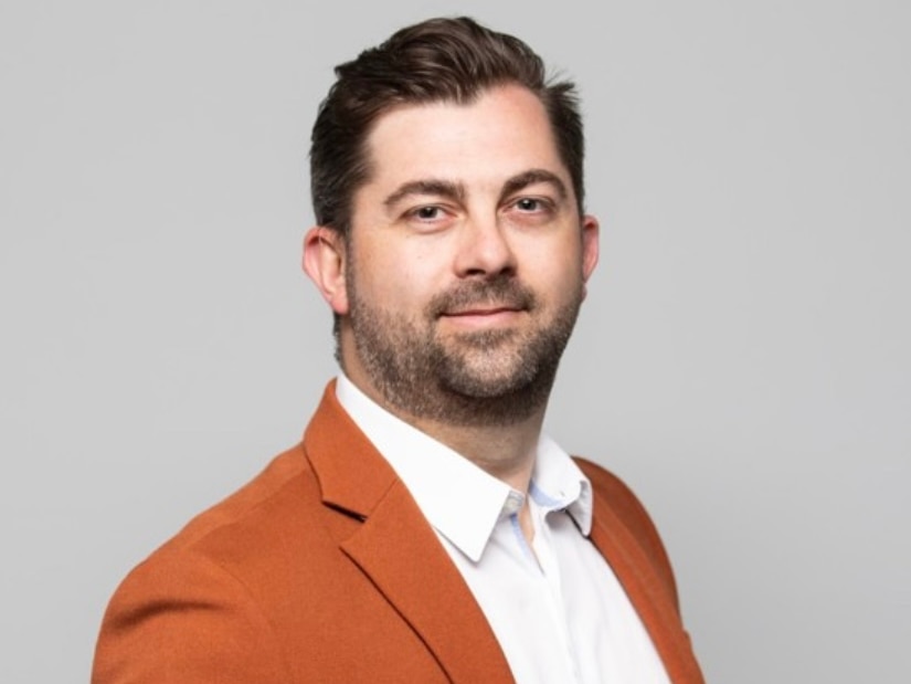 A photo of a man with brown hair and a beard, wearing a white collared shirt and a light orange blazer, grey background