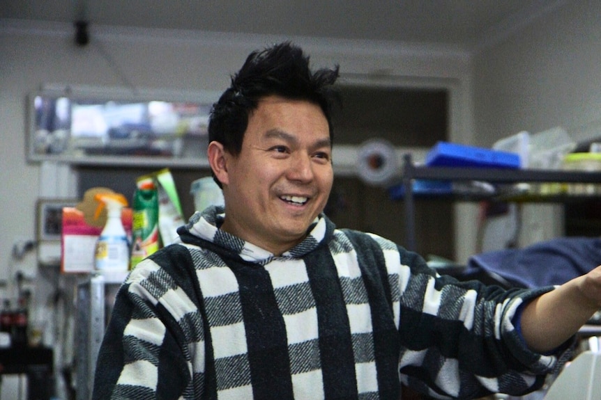 A man with an Asian face wearing a black and white checked shirt gestures ahead with industrial kitchen shelves behind