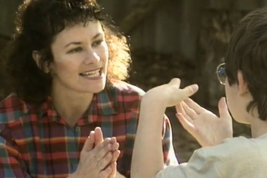 Woman with curly dark hair and wearing a red and green check blouse mid hand clap with her son who sits across from her