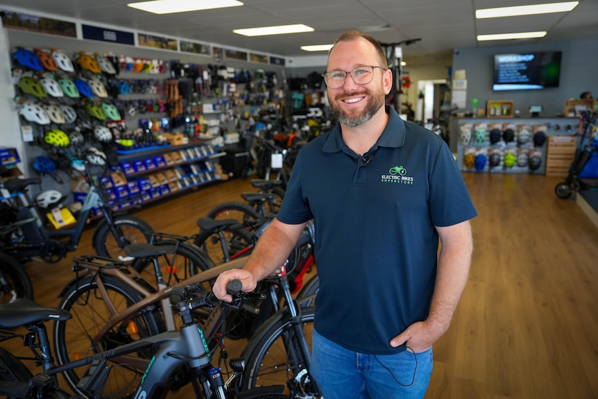 A man has a beard, glasses and a navy polo shirt, he stands leaning on a bike in a store with cycling accessories.