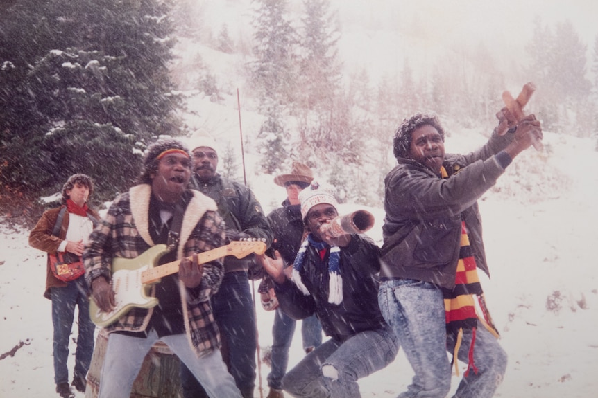 Six men in winter beanies and jackets look joyous in heavy snowfall. One man in the front is holding an electric guitar.